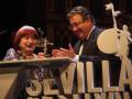A una mujer y un hombre en un escenario, posiblemente durante un evento cultural o artístico. La mujer sostiene un trofeo mientras el hombre la felicita con una sonrisa. La escena se desarrolla en un entorno que parece ser el Festival de Cine de Sevilla, como indica la inscripción en el fondo.
