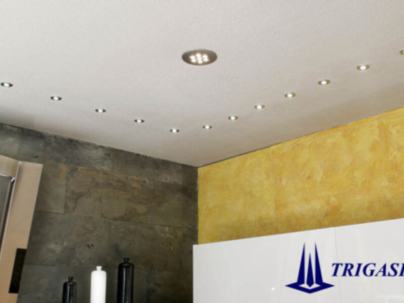 Interior de una oficina con techo de iluminación LED y paredes de textura amarilla con logo de Trigasia en primer plano.