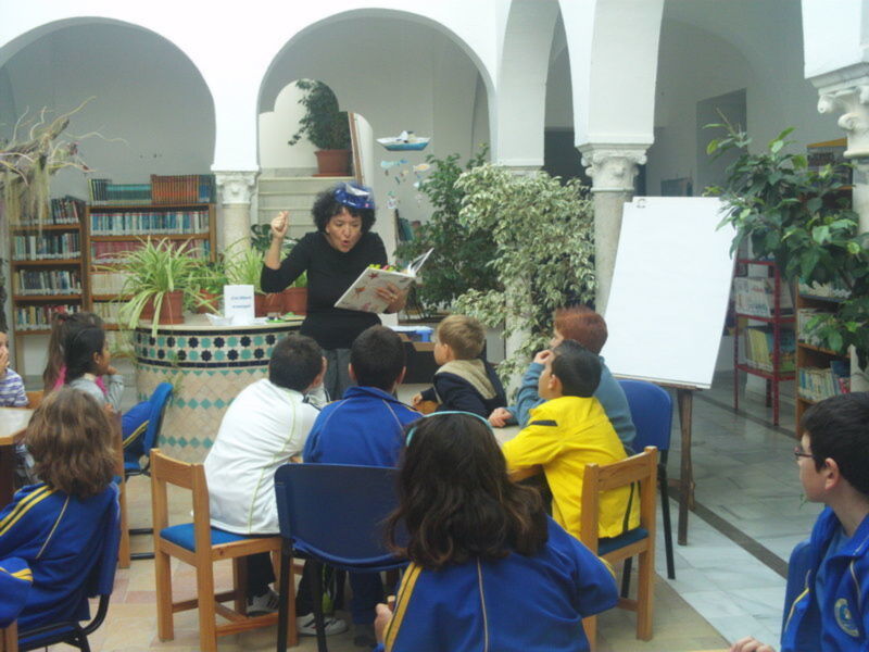 Profesor leyendo a los niños en una biblioteca con plantas y libros en el fondo.