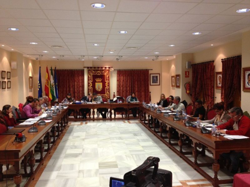 Sala de reuniones con mesas largas y sillas, personas sentadas, banderas y un escudo en el fondo.