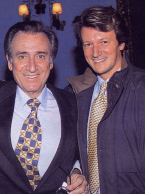 Dos hombres sonrientes en trajes, posando juntos en un ambiente formal.