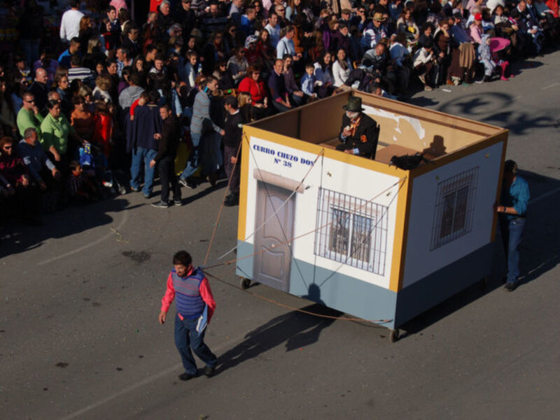 Desfile con carroza temática en una celebración pública, con un hombre en un pequeño carroza representando una casa y un espectador en el fondo.