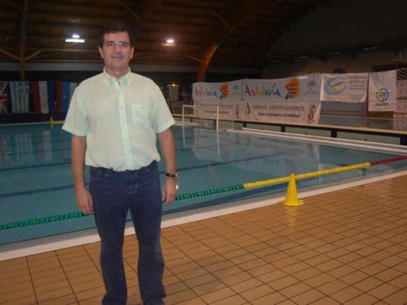 Un hombre posa frente a una piscina con banderas de diferentes países en el fondo.