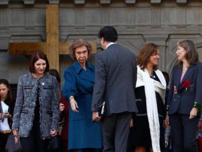 Una mujer de edad avanzada camina entre una fila de personas en una reunión formal, rodeada por una arquitectura histórica.