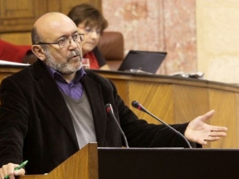 Persona hablando en un foro o reunión en un ambiente formal, con un micrófono y un panel de fondo.