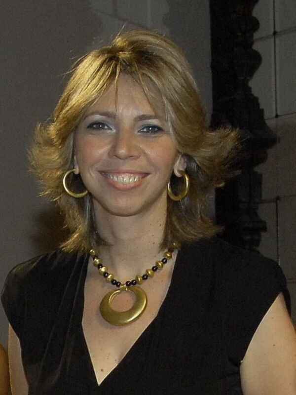 Retrato de mujer sonriente con cabello rubio y joyas doradas, vestida con un top negro y collar circular.