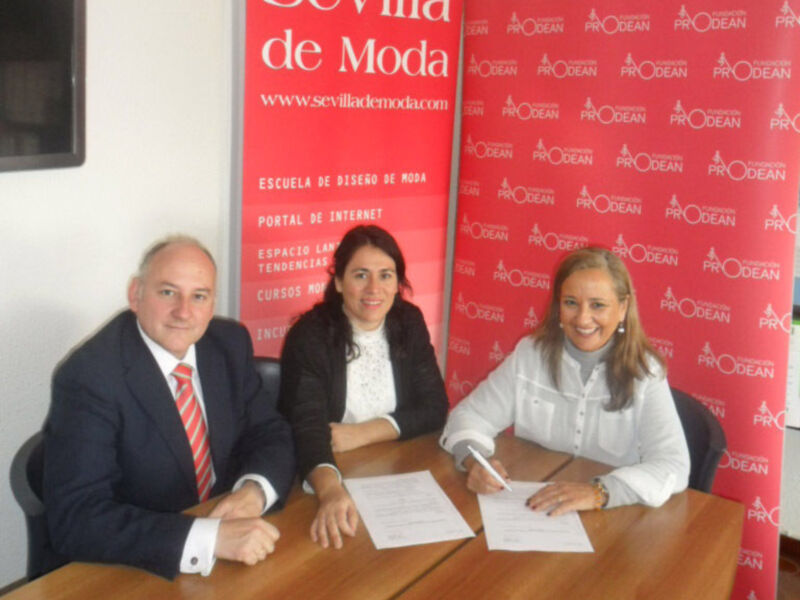 Tres personas firmando documentos en una mesa frente a un cartel de Sevilla de Moda.
