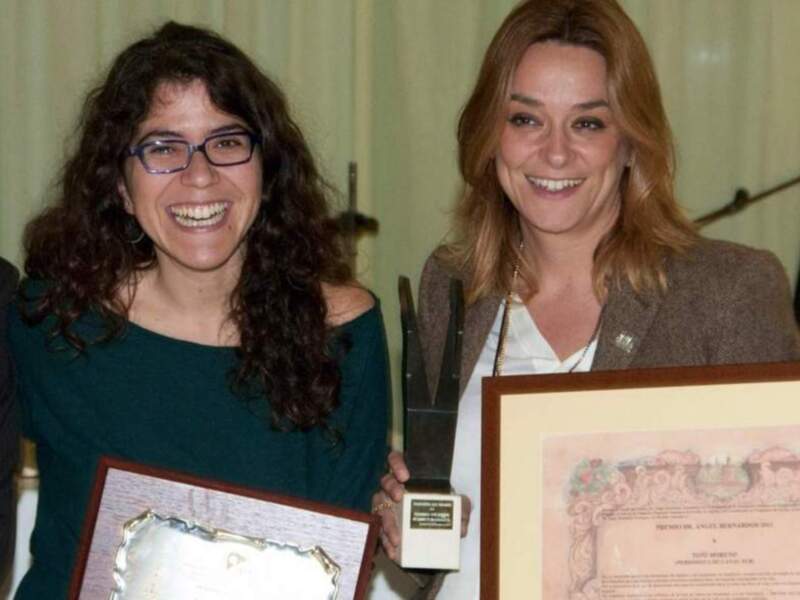 Dos mujeres sonríen mientras reciben un premio y un diploma.