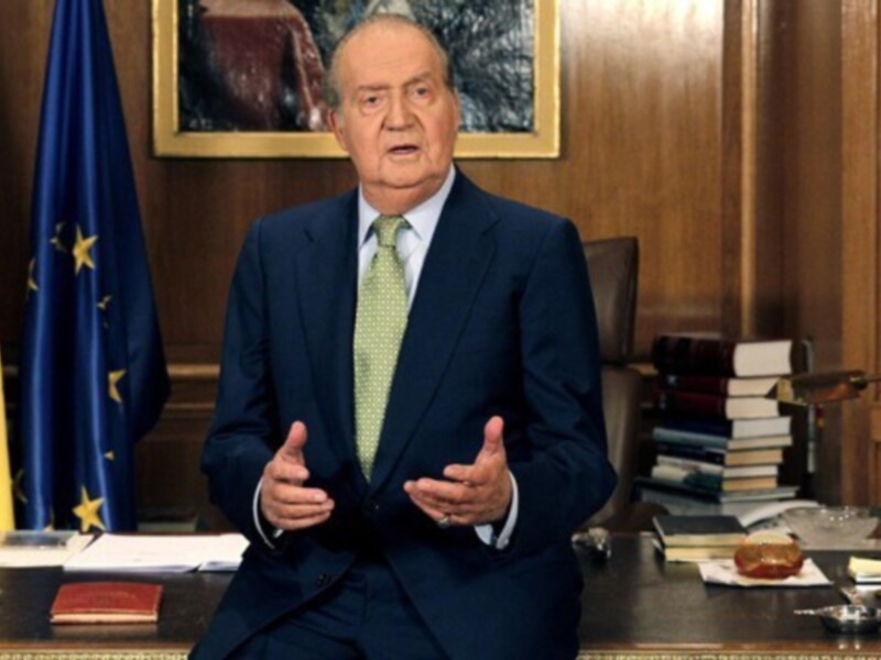 El rey Juan Carlos I de España en su despacho, rodeado de símbolos de la monarquía española y la Unión Europea.