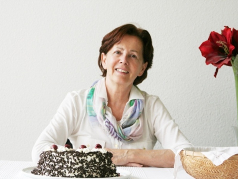 Una mujer sonríe mientras disfruta de un pastel de chocolate con crema y frutos rojos en una mesa decorada con una flor roja.