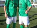 Dos hombres en camisetas verdes y pantalones blancos posan juntos en un campo de fútbol.