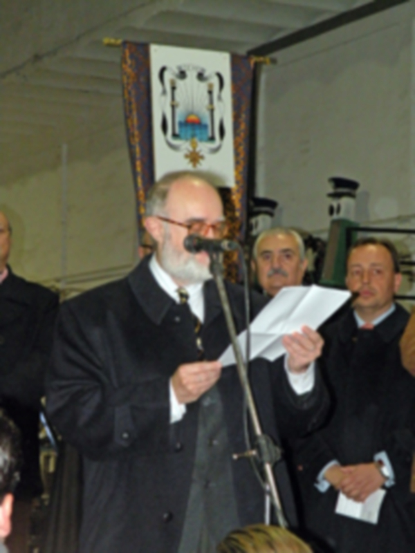 Persona en traje formal leyendo un documento en un evento público con bandera en el fondo.