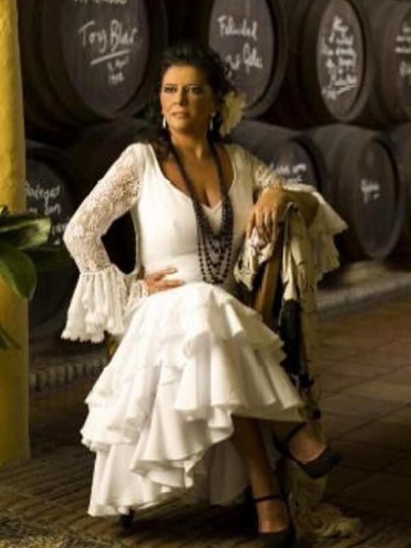Una mujer vestida con un vestido blanco con volantes y una chaqueta beige, posando frente a barriles de vino en un entorno de bodega.
