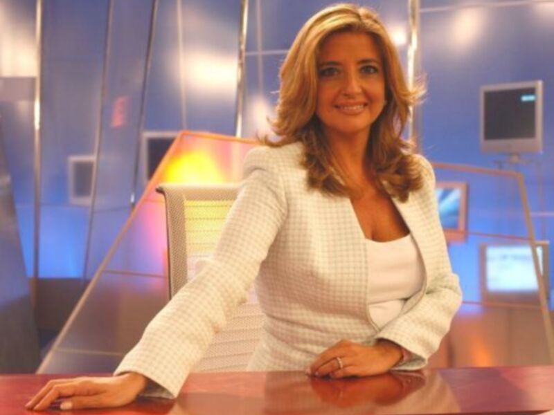 Una mujer con cabello largo y rizado, vistiendo una chaqueta blanca, posa con las manos en la mesa en un entorno de estudio.