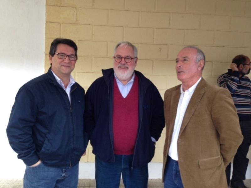 Tres personas sonrientes, dos de ellas con chaquetas y una con una camisa blanca, posando frente a una pared de ladrillos amarillos.