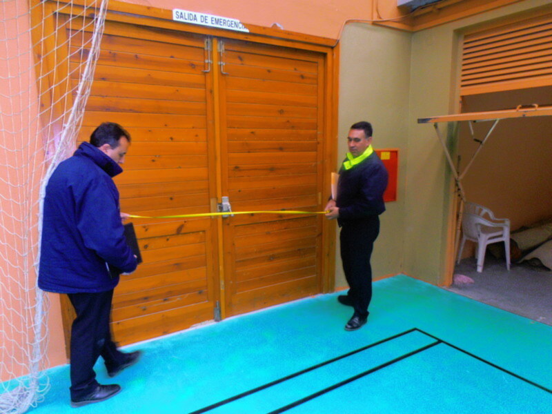 Dos hombres frente a una puerta de madera con un cartel que dice 'SALIDA DE EMERGENCIA'. Uno de ellos sostiene un cinta de corte amarilla. El fondo muestra un área interior con una silla blanca y una pared de color naranja.