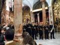 Una banda militar toca en un ambiente histórico con columnas y arcos dorados, mientras una multitud observa.