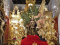Imagen de un altar dorado con una figura central vestida en negro y rodeada por ángeles dorados. La imagen está iluminada con luces doradas y un fondo de madera tallada, creando una atmósfera religiosa y sagrada.