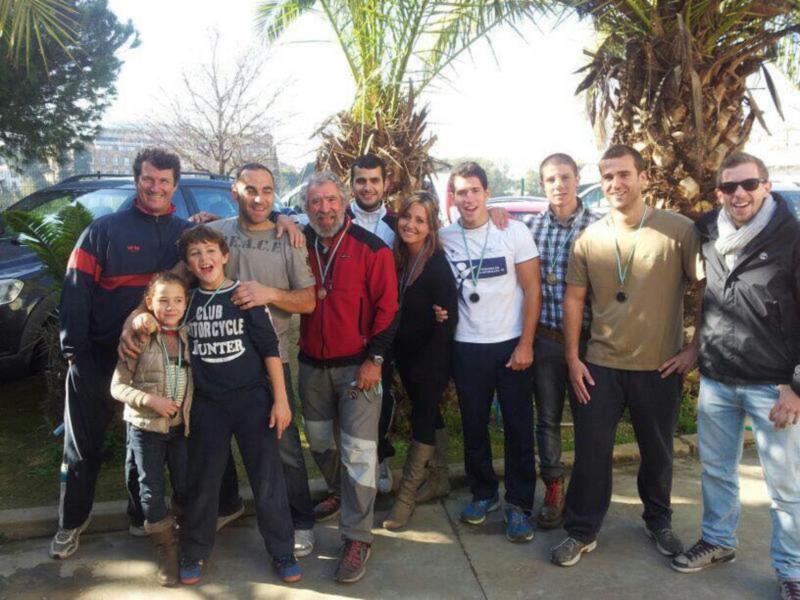 Un grupo de personas, incluyendo niños y adultos, sonríe y posa juntos en un día soleado al aire libre, rodeados de árboles y coches.