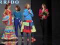Desfile de moda con modelos en trajes coloridos y una presentadora con micrófono en escenario.