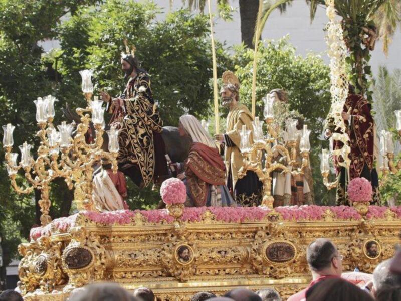 Procesión religiosa con imágenes procesionales adornadas con flores y candelabros dorados, celebrada en un entorno urbano con árboles y palmeras al fondo.