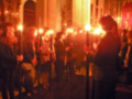 Una procesión religiosa con personas vestidas de negro y rojo, sostienen antorchas encendidas en una iglesia con arcos y vitrales.