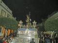 Procesión nocturna con una imagen religiosa en el centro, rodeada de personas y luces festivas en un entorno urbano con edificios y árboles alrededor.