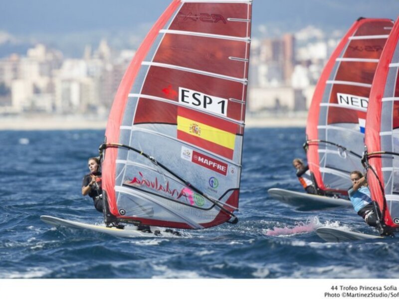 Vela de windsurf con la bandera española y la marca Mapfre en un evento de vela en el mar Mediterráneo, con un paisaje urbano en el fondo.