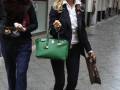 Dos mujeres caminando por una calle adoquinada, una lleva un bolso verde y la otra un bolso negro, con ropa elegante y moderna.