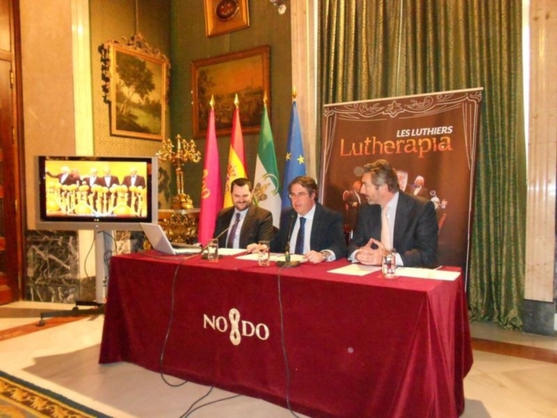 Se puede ver a una conferencia de prensa en un salón elegante con tapices y cortinas verdes. En el centro, hay una mesa cubierta con un mantel rojo y la bandera de España. Los asistentes están sentados en una mesa con un monitor en el fondo que muestra a personas. En la pared hay dos cuadros y una lámpara de araña. La imagen también incluye un cartel con la palabra "Lutherapia" en el fondo.