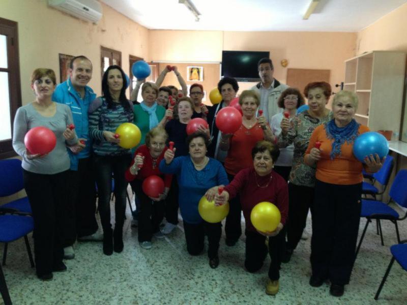 Un grupo de personas, incluyendo adultos mayores, participando en una actividad de ejercicios con balones de colores en una sala de conferencias. La diversión y la actividad física son evidentes en la expresión y el gesto de los participantes.