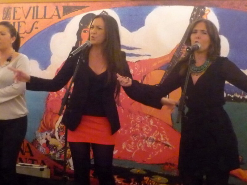 Tres mujeres cantando en un escenario con un fondo decorativo de Sevilla.