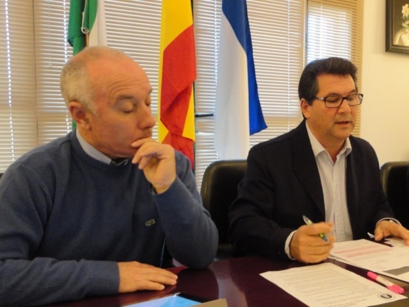 Dos hombres en una reunión formal, uno escribiendo en un documento mientras habla.