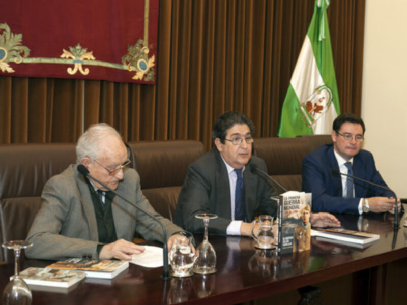 Tres hombres sentados en una mesa con libros y copas de vino, con una bandera y una bandera nacional en el fondo.