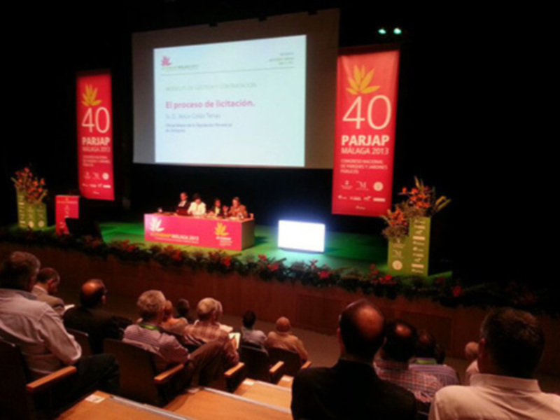 Una sala de conferencias con una pantalla grande mostrando un documento con el título 'El proceso de licitación' y el logo de 'PARIAP'. La sala está decorada con flores y hay personas sentadas en los asientos.