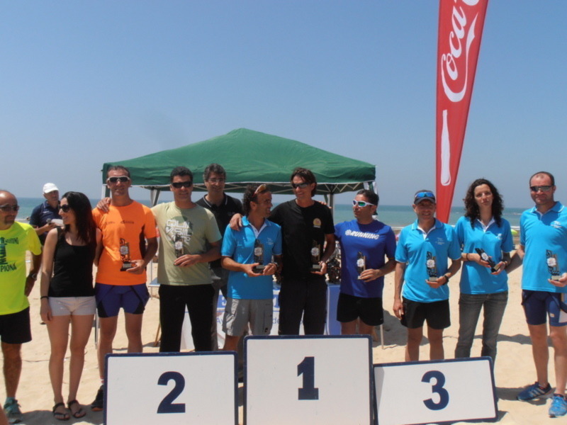 Equipo de atletas celebrando en el podio de una competición de atletismo en la playa, con trofeos y bandera de Coca-Cola al fondo.