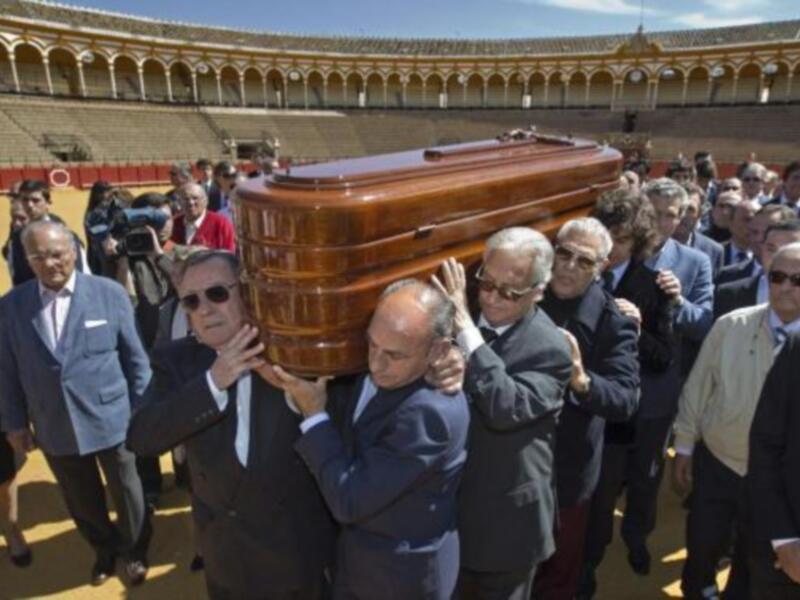 Un grupo de personas portando un ataúd en un entorno de plaza de toros, rodeado por espectadores.