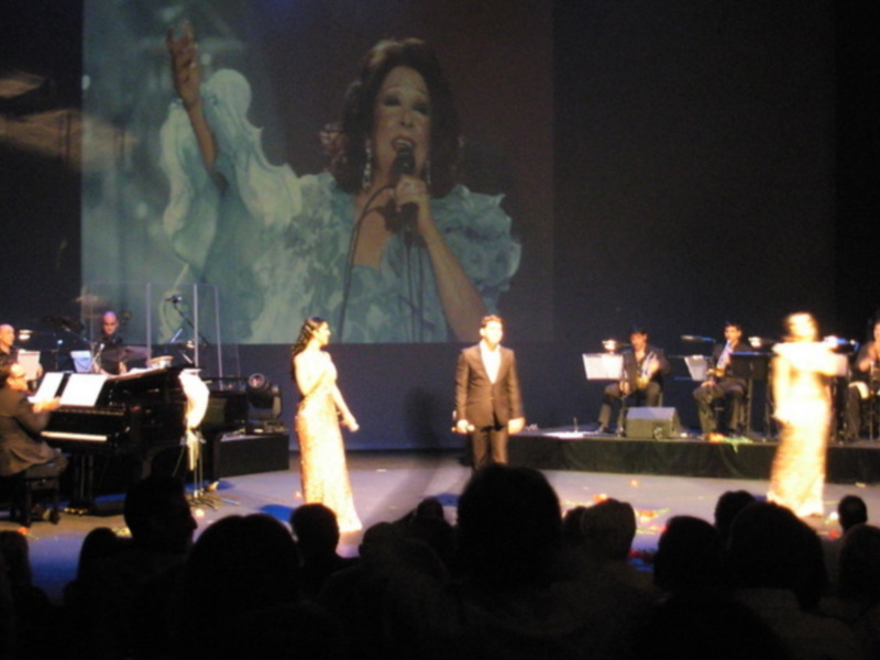 Una cantante en escenario con un piano y un coro en el fondo, iluminada por luces de escenario.