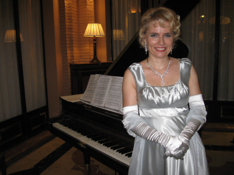 Elegant woman in a silver dress and gloves standing beside a grand piano, ready to perform.