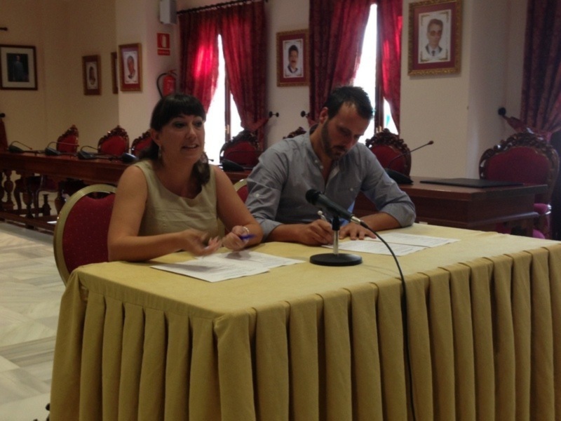 Dos personas sentadas en una sala de conferencias, uno escribiendo y la otra hablando, con un micro en el escritorio y varias fotos en la pared al fondo.