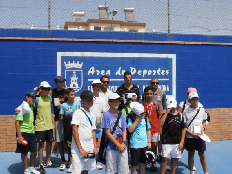 Grupo de personas posando en un campo de tenis con una pared azul y una bandera de España en el fondo.