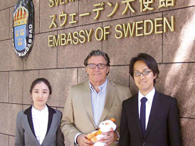 Tres personas posan frente a la Embajada de Suecia en Japón, con un hombre en el centro sosteniendo un peluche y dos personas a su izquierda y derecha, uno de ellos sonriendo ampliamente.