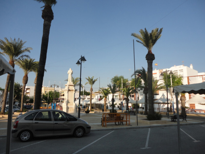 Plaza con palmeras y estatua en un día soleado, con edificios blancos al fondo.