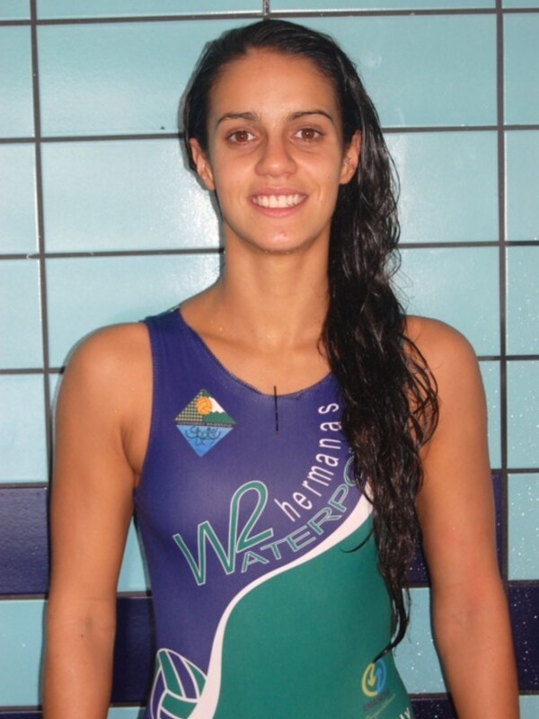 Una mujer con una camiseta azul y verde con el logo de 'WATERPOLO HERMANAS' posando frente a una pared azul con cuadros.