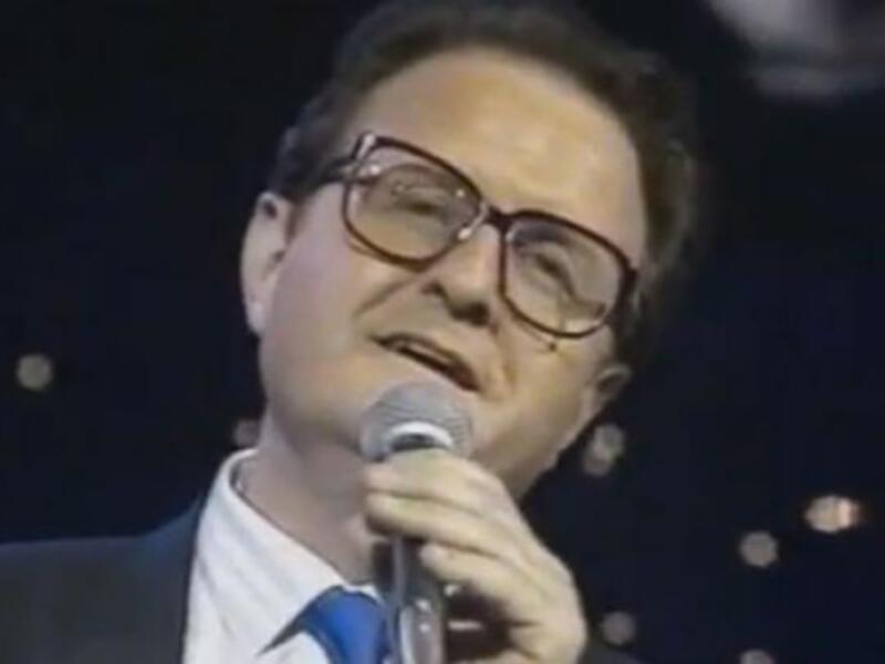 Persona cantando en un escenario con un micrófono, vestida con un traje y corbata, en un ambiente de concierto o gala.
