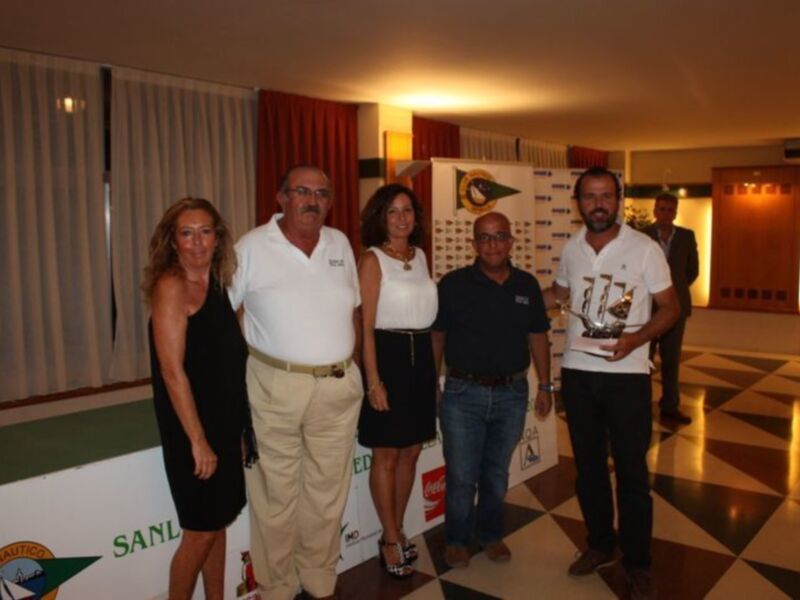 Equipo de negocios posando con un premio en un evento corporativo. El ambiente es formal, con vestimenta elegante y un fondo con logotipos de empresas. La iluminación es suave y cálida, creando un ambiente acogedor.