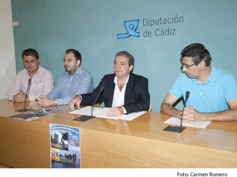 Cuatro hombres sentados en una mesa con folletos y un micro en frente de ellos, con el logotipo de la Diputación de Cádiz en el fondo.