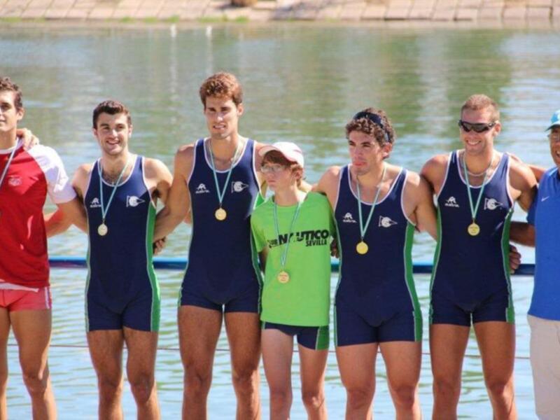 Equipo de remo celebrando con medallas en un lago, con un entrenador en la última posición. Todos están sonriendo y parecen estar muy satisfechos con su logro.