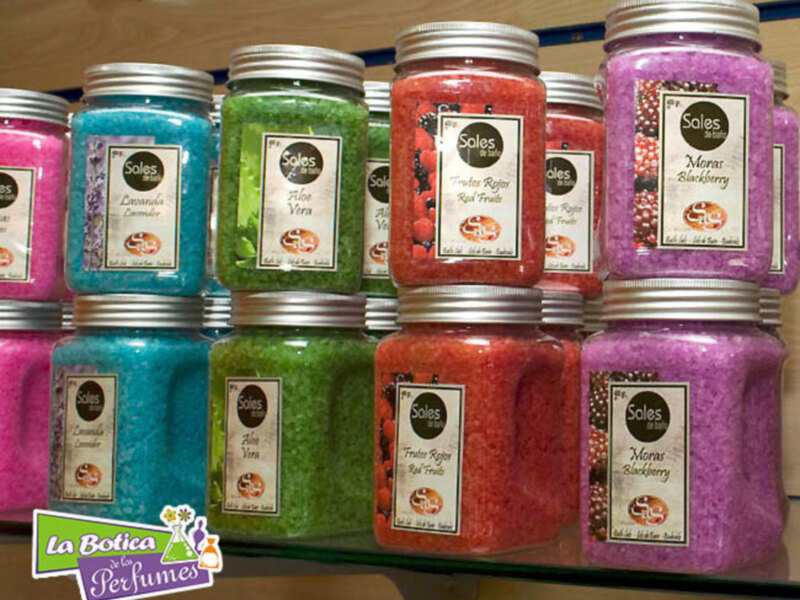 Varios frascos de sales de baño de colores y sabores, como Aloe Vera y Moras, en una estantería.