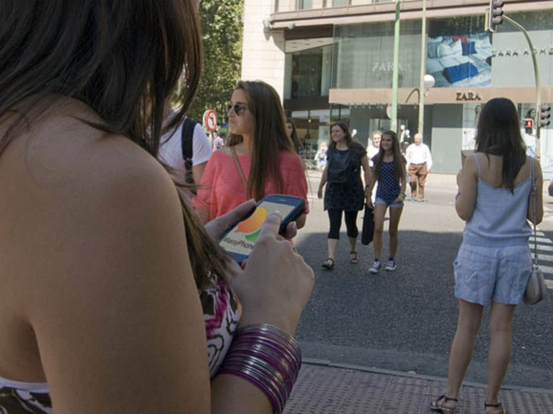 Una mujer sostiene un teléfono móvil en una calle con tiendas y personas a pie.